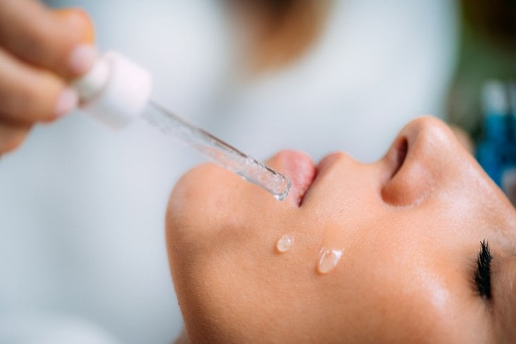Einer Frau mit geschlossenen Augen werden zwei Hyaluronsäure-Serum-Tropfen auf die Wange geträufelt